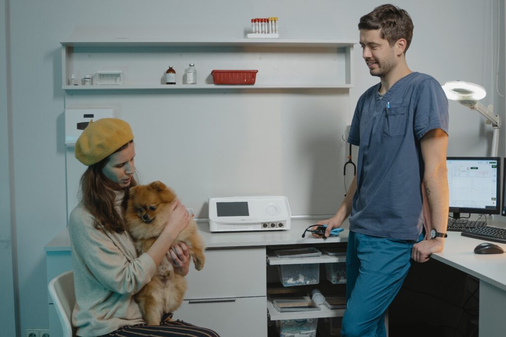 A woman holding her pet dog is sitting on a chair, in a vet while the vet is looking at her and the pet dog. 
Tips for taking care of a pet dog include taaking your pet dog for regular vet vsists.